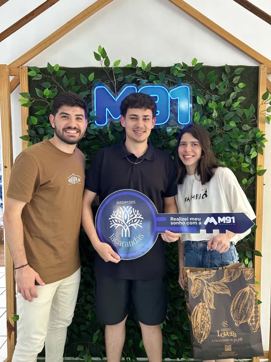 Trio diante de painel decorado com folhagem, o rapaz do meio segura uma placa comemorativa em formato de chave; a mulher à direita carrega uma sacola de presente de chocolateria.