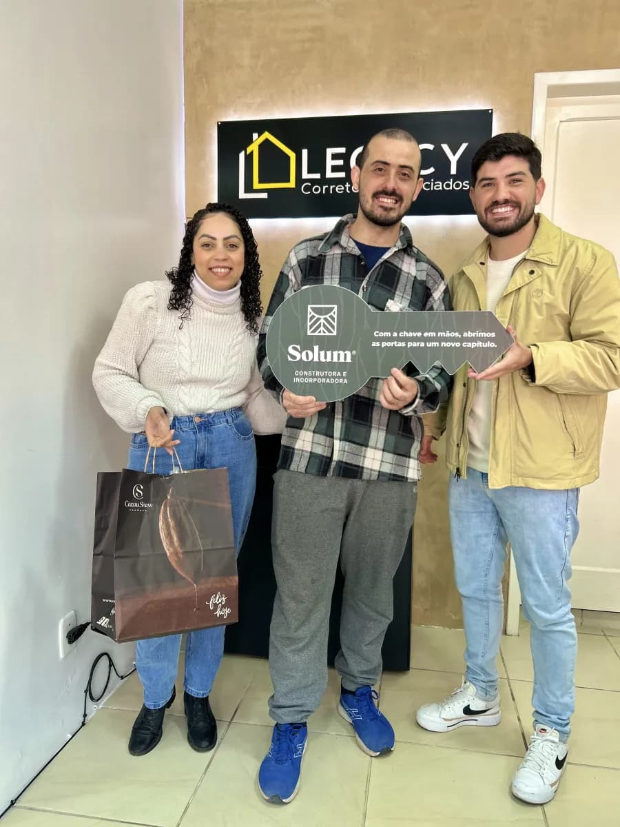 Casal com Matheos diante do painel de uma imobiliária, o homem do meio segura uma placa comemorativa em formato de chave; a mulher carrega uma sacola de presente de chocolateria.