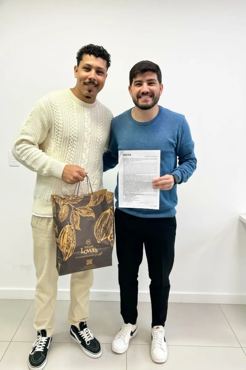 Cliente ao lado de Matheos em ambiente interno claro, Matheos segura o contrato da Caixa recém-assinado; o cliente carrega uma sacola de presente de chocolateria.