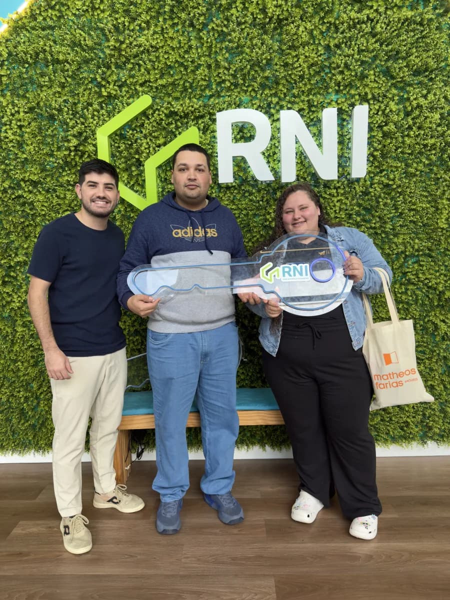 Casal com Matheos diante de painel verde de folhagem, a mulher segura uma chave cerimonial de acrílico e carrega uma sacola de tecido com a marca da imobiliária.
