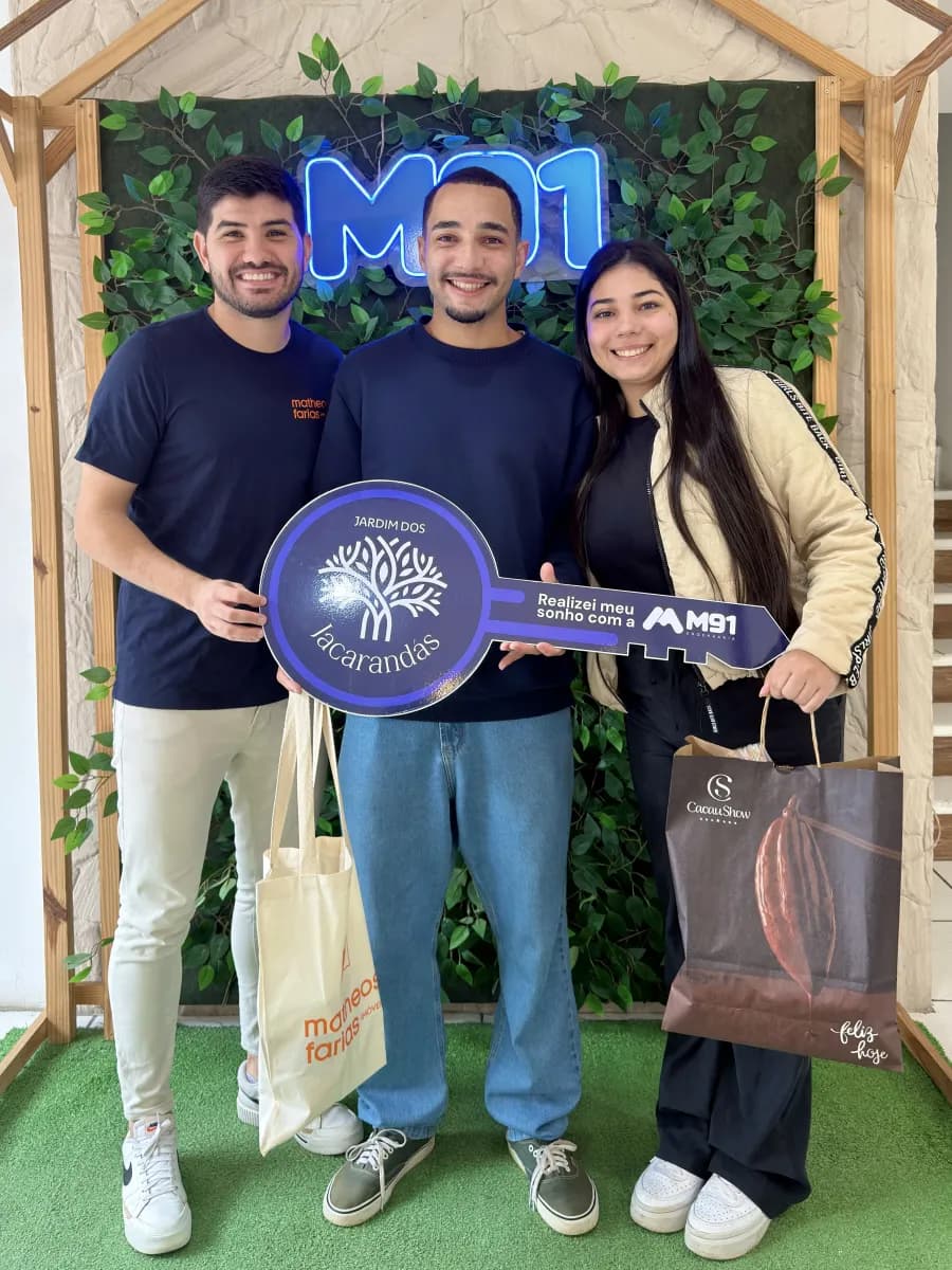 Trio diante de painel decorado com folhagem sobre grama sintética, o rapaz do meio segura uma placa comemorativa em formato de chave; a moça à direita carrega uma sacola de presente de chocolateria.