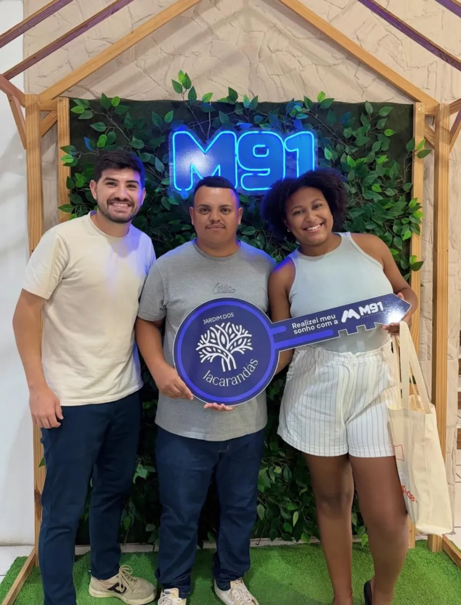 Trio diante de painel decorado com folhagem sobre grama sintética, o rapaz do meio segura uma placa comemorativa em formato de chave; a moça à direita, de cabelo cacheado, carrega uma sacola de tecido.