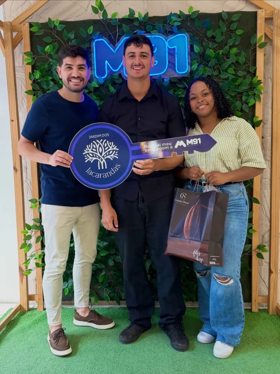 Trio diante de painel decorado com folhagem sobre grama sintética, o rapaz de camisa escura segura uma placa comemorativa em formato de chave; a moça à direita carrega uma sacola de presente de chocolateria.