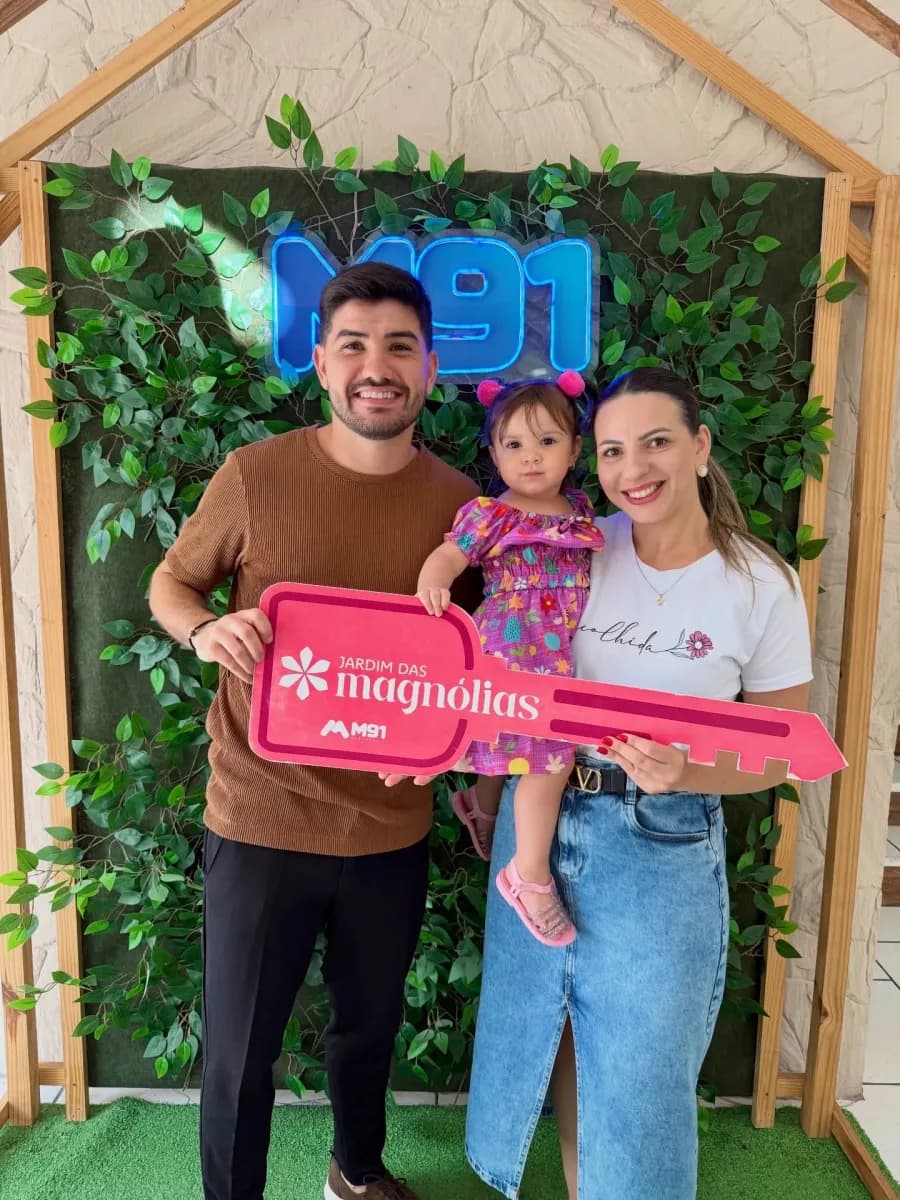 Casal com uma criança pequena no colo diante de painel decorado com folhagem, segurando juntos uma placa comemorativa cor-de-rosa em formato de chave.