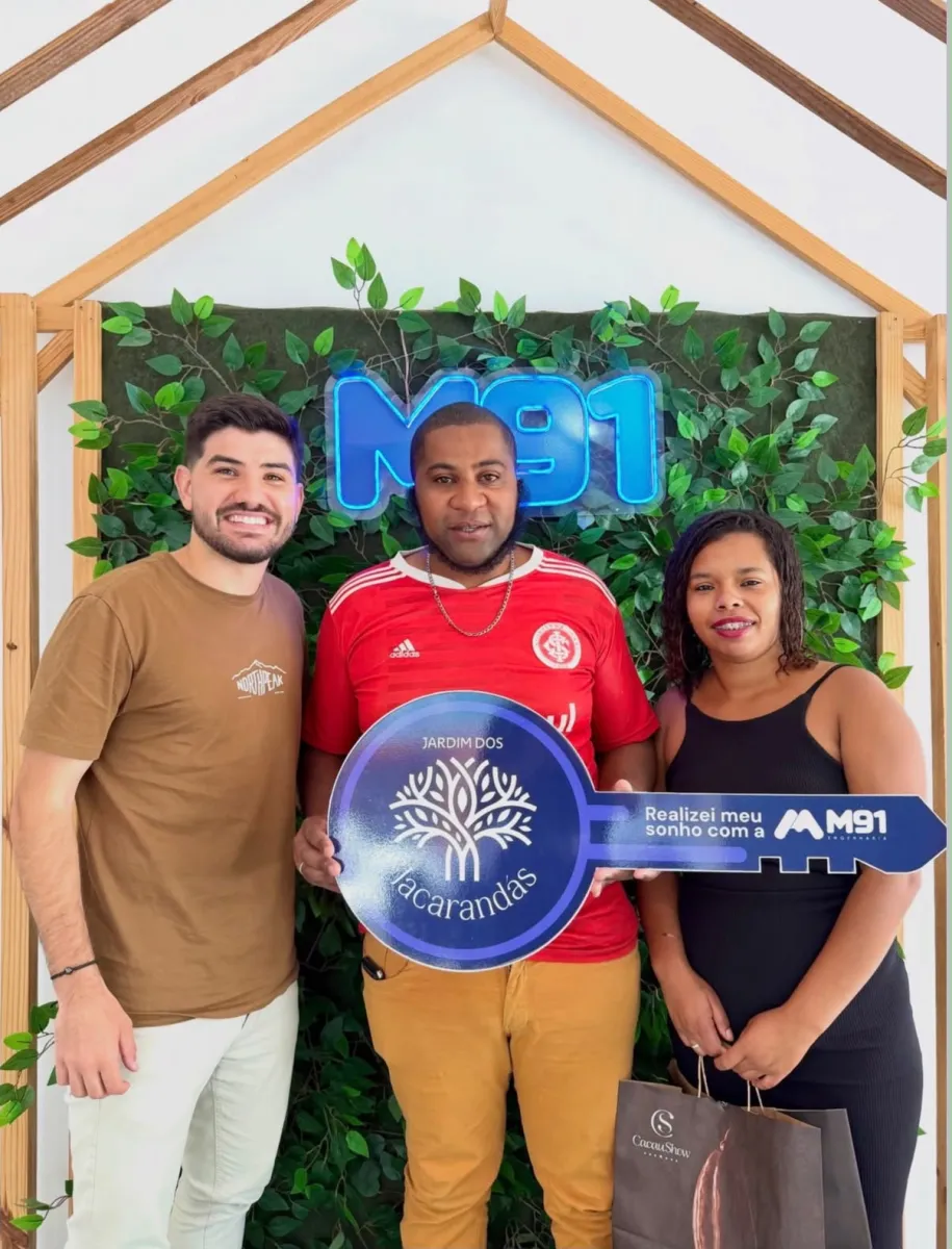 Trio sorridente diante de painel decorado com folhagem, o homem do meio veste camisa de time de futebol e segura uma placa comemorativa em formato de chave; a mulher à direita carrega uma sacola de presente de chocolateria.