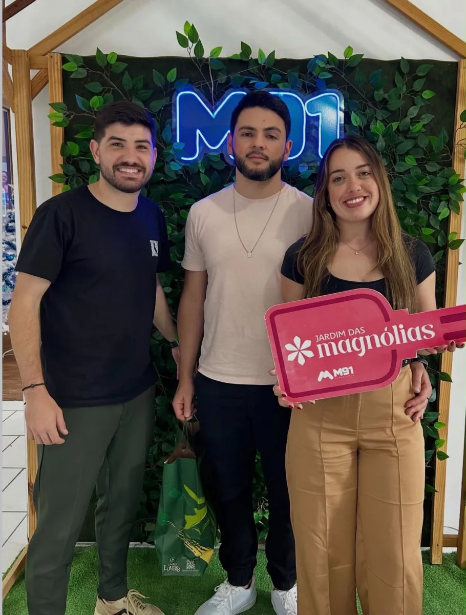 Trio diante de painel decorado com folhagem; a mulher à direita segura uma placa comemorativa cor-de-rosa em formato de chave, e o homem do meio carrega uma sacola verde de presente ao seu lado.