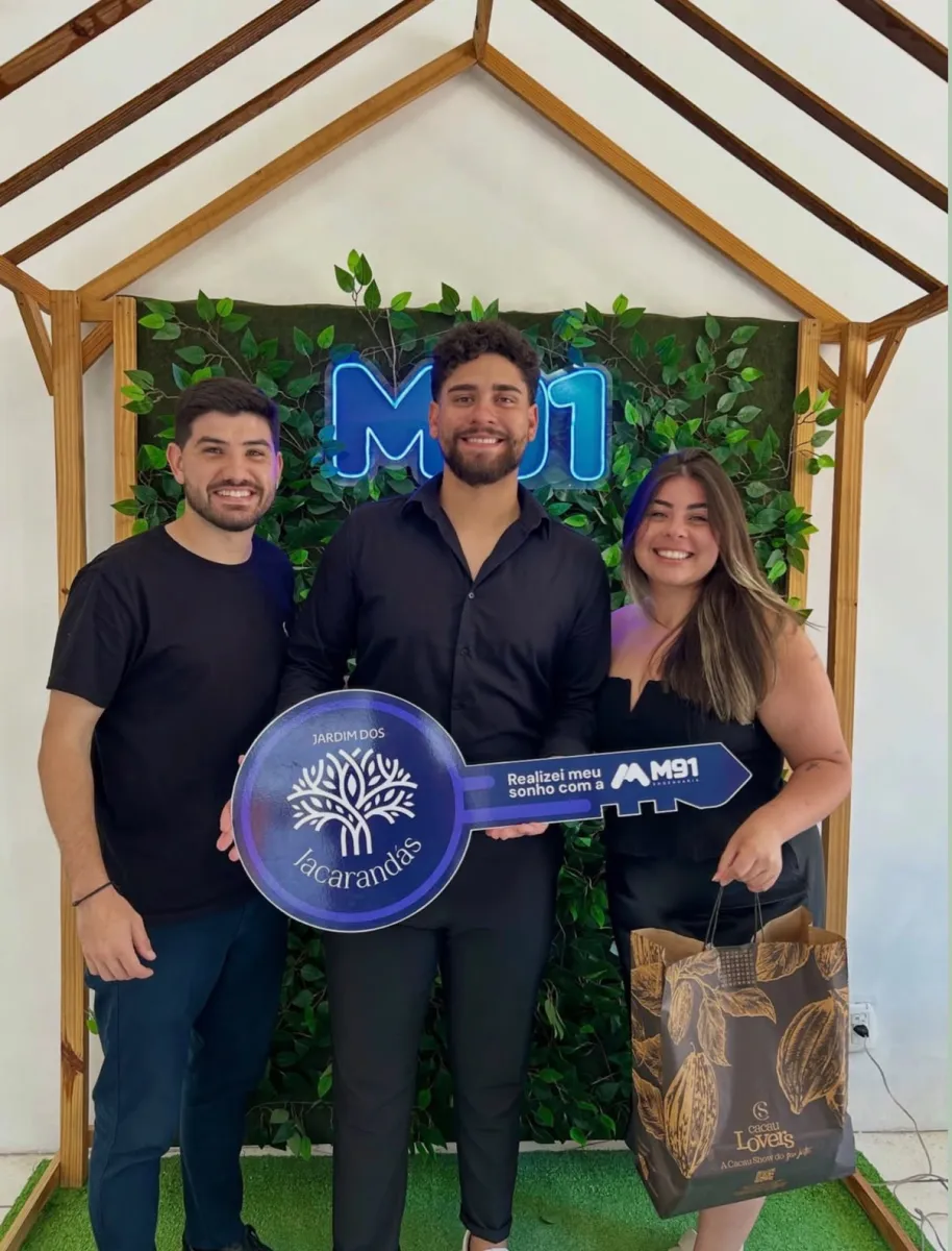 Trio diante de painel decorado com folhagem, o homem do meio segura uma placa comemorativa em formato de chave; a mulher à direita, de cabelo com mechas claras, sorri ao lado dele.