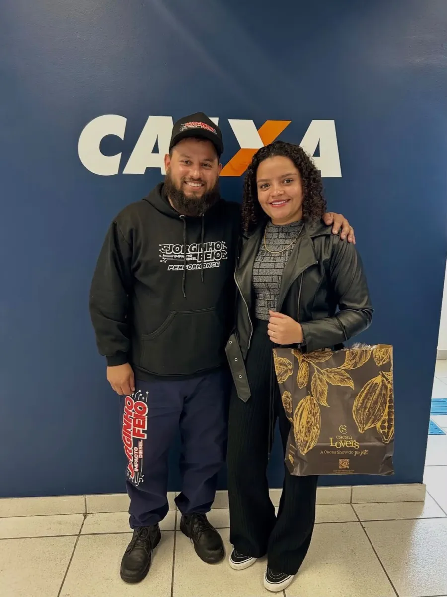 Casal em frente a uma parede com o logotipo da Caixa, o homem veste boné e moletom com estampa automotiva; a mulher, de cabelo cacheado, carrega uma sacola de presente de chocolateria.