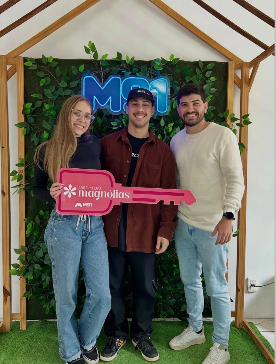 Trio diante de painel decorado com folhagem, a mulher à esquerda e o rapaz de boné no meio seguram juntos uma placa comemorativa cor-de-rosa em formato de chave.