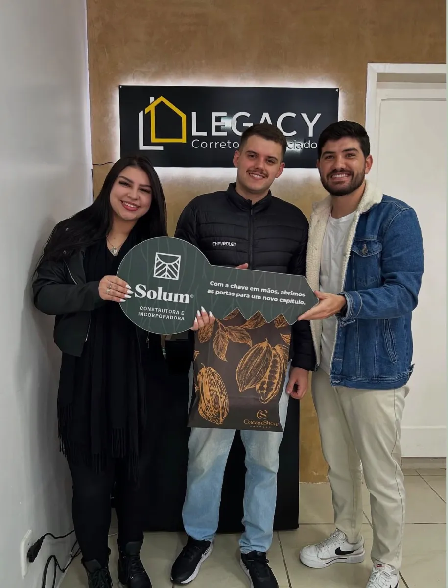 Trio diante do painel de uma imobiliária; a mulher à esquerda e o homem de jaqueta jeans à direita seguram juntos uma placa comemorativa em formato de chave, enquanto o homem do meio, de jaqueta escura, não a toca.